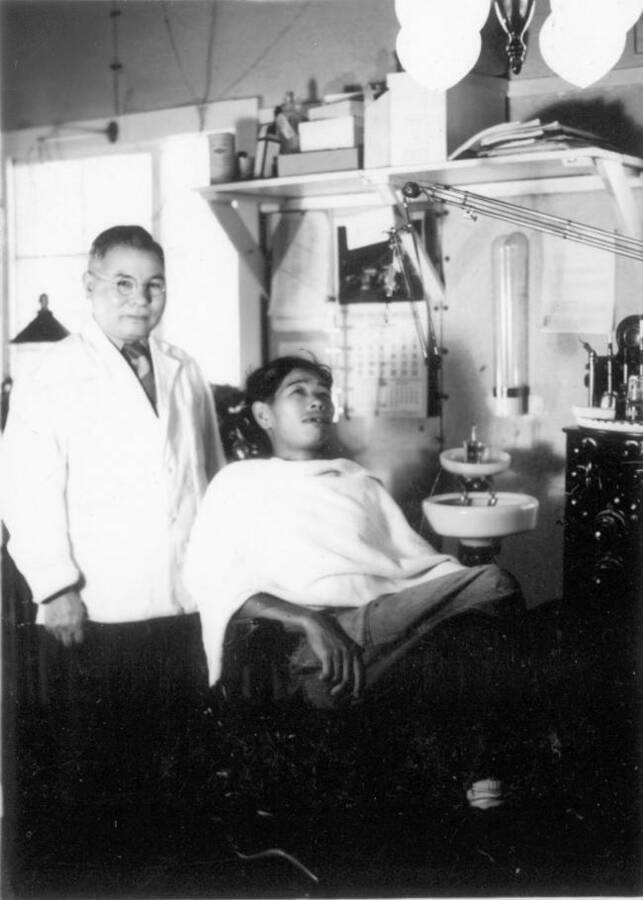 Interior image of two men: one standing wearing a medical jacket, while other sits in dentist chair at Kooskia Internment Camp. Photo taken from 12-3/4 x 15-1/4 Photograph album of the Kooskia Japanese Internment Camp.