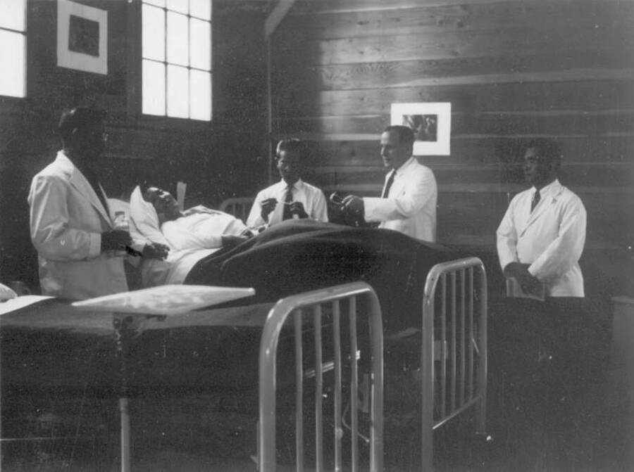 Interior image of men in medical jackets attending a patient in bed at Kooskia Internment Camp. Photo taken from 12-3/4 x 15-1/4 Photograph album of the Kooskia Japanese Internment Camp.