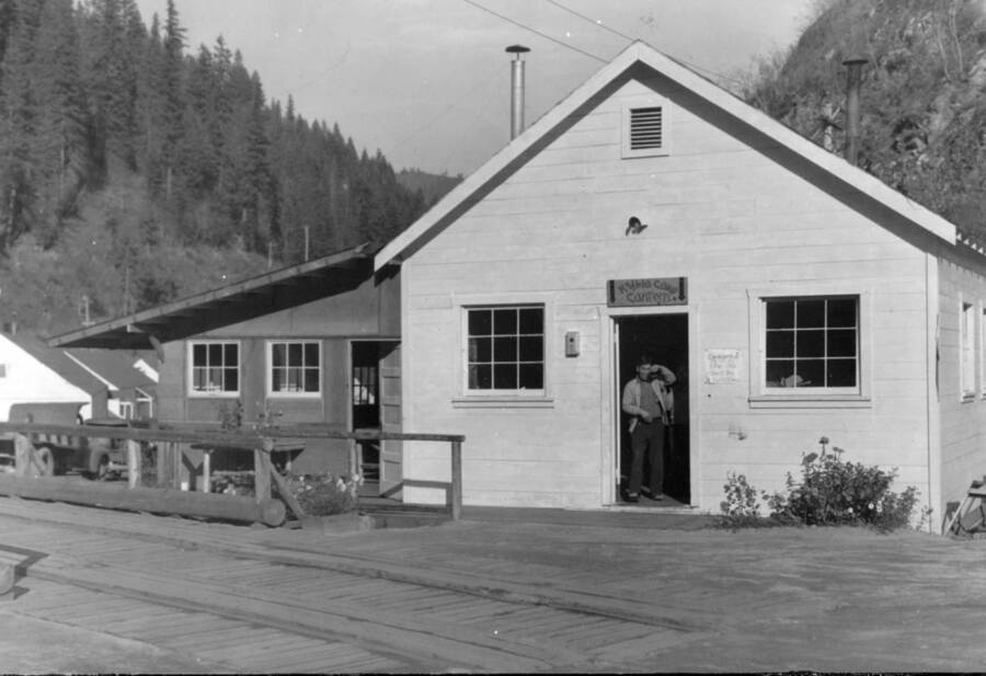 Image of buildings with a man in a doorway; sign on buildings says, "Kooskia Camp Canteen". Photo taken from 12-3/4 x 15-1/4 Photograph album of the Kooskia Japanese Internment Camp.