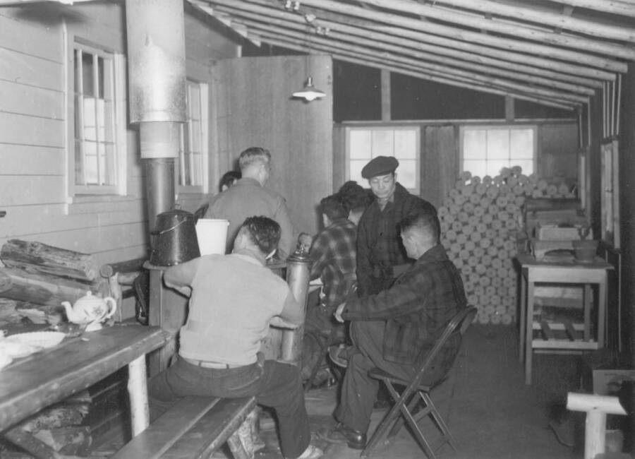 Interior image of a group of men sitting around a wood-burning stove at Kooskia Internment Camp. Photo taken from 12-3/4 x 15-1/4 Photograph album of the Kooskia Japanese Internment Camp.