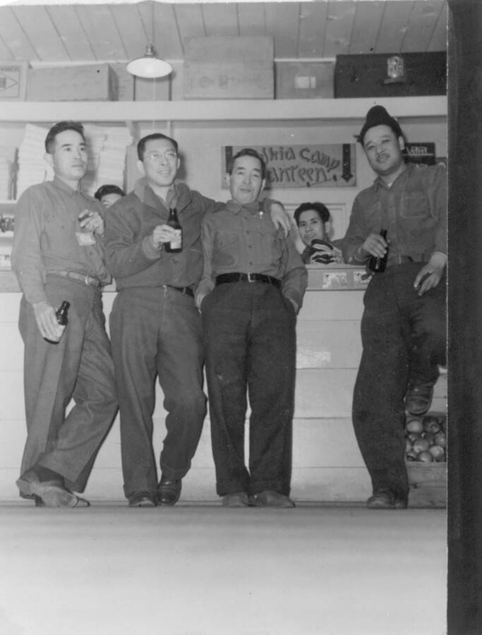 Interior view of group of  men in front of store counter drinking beverages; sign behind counter reads, "Kooskia Camp Canteen". Photo taken from 12-3/4 x 15-1/4 Photograph album of the Kooskia Japanese Internment Camp.
