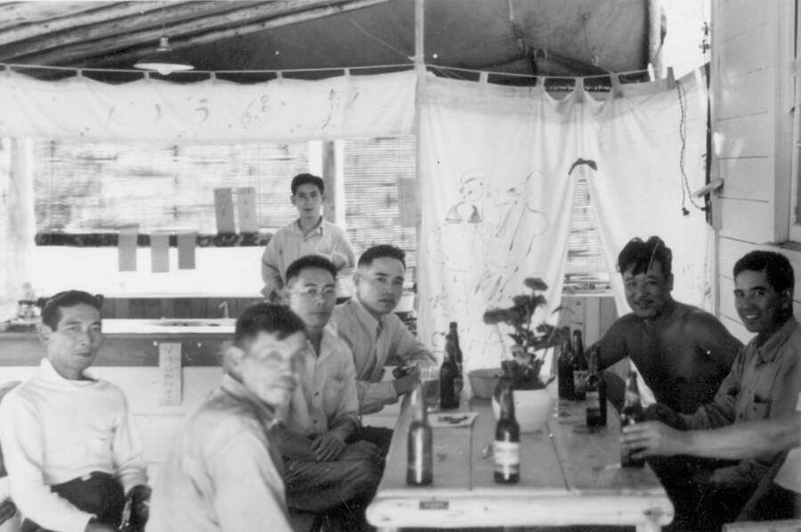 Image of a group of men sitting at a table drinking bottled beverages outside the canteen at Kooskia Internment Camp. Photo taken from 12-3/4 x 15-1/4 Photograph album of the Kooskia Japanese Internment Camp.