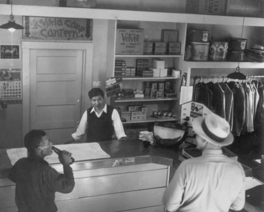 Interior image of three men in store talking; sign in background says, "Kooskia Camp Canteen". Photo taken from 12-3/4 x 15-1/4 Photograph album of the Kooskia Japanese Internment Camp.