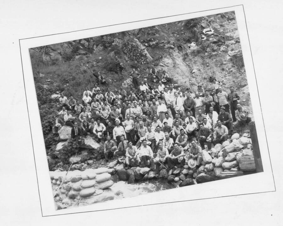 A group photograph of the workers of the Kooskia Internment Camp beside the Lochsa River. Sandbags can be seen in the foreground alongside the river. Photo taken from 12-3/4 x 15-1/4 Photograph album of the Kooskia Japanese Internment Camp.