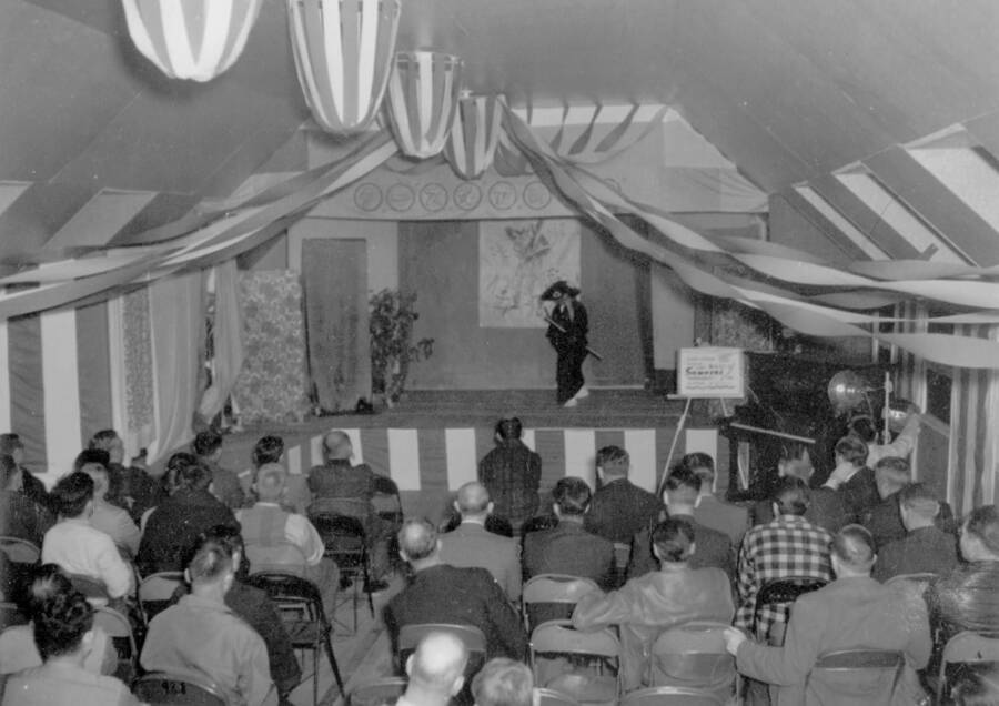 Interior image of a stage performance by a man in traditional costume at Kooskia Internment Camp. Photo taken from 12-3/4 x 15-1/4 Photograph album of the Kooskia Japanese Internment Camp.