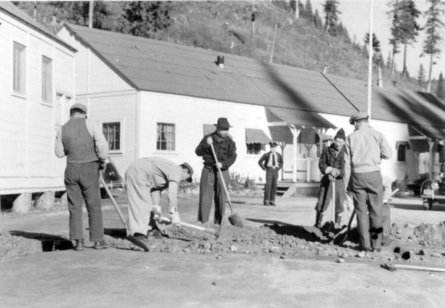 Image of men at Kooskia Internment Camp digging with shovels and pickaxes; an officer (?) is standing in background. Photo taken from 12-3/4 x 15-1/4 Photograph album of the Kooskia Japanese Internment Camp.