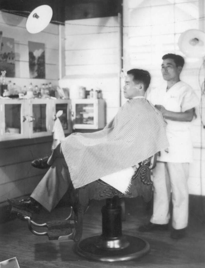 Interior image of two men: one is  sitting in barber chair at Kooskia Internment Camp. Photo taken from 12-3/4 x 15-1/4 Photograph album of the Kooskia Japanese Internment Camp.