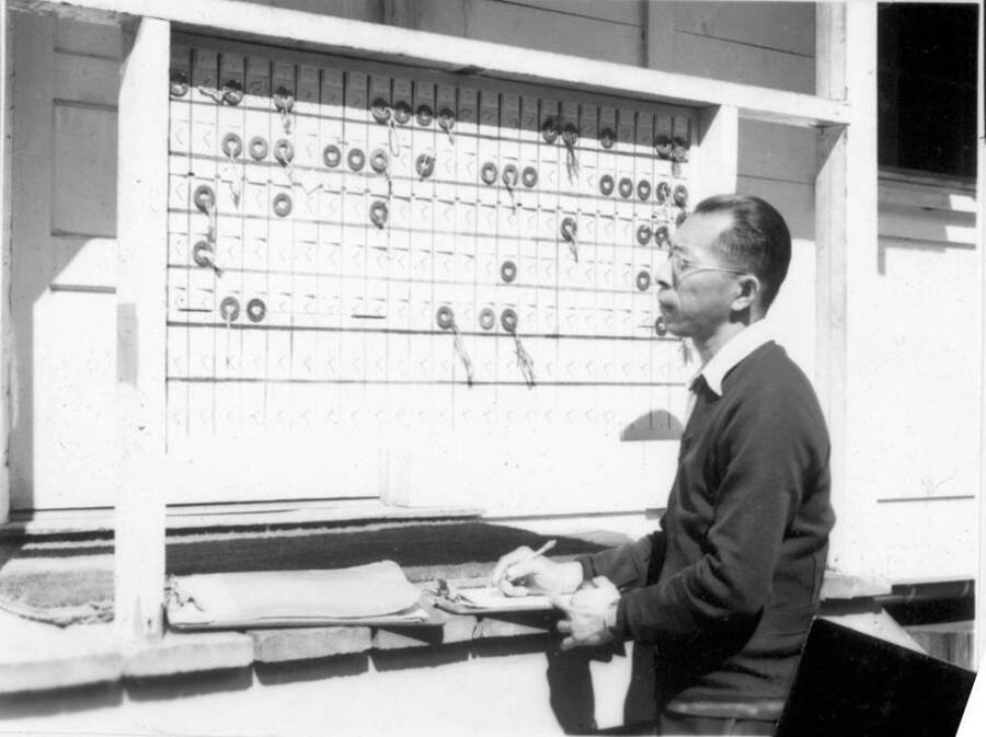 Image of a man with a clipboard in front of a check-in board. Photo taken from 12-3/4 x 15-1/4 Photograph album of the Kooskia Japanese Internment Camp.