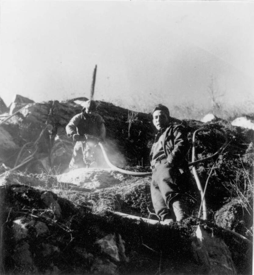Image of two men working on a construction project on hillside at Kooskia Internment Camp; one is pictured using a jackhammer. Photo taken from 12-3/4 x 15-1/4 Photograph album of the Kooskia Japanese Internment Camp.