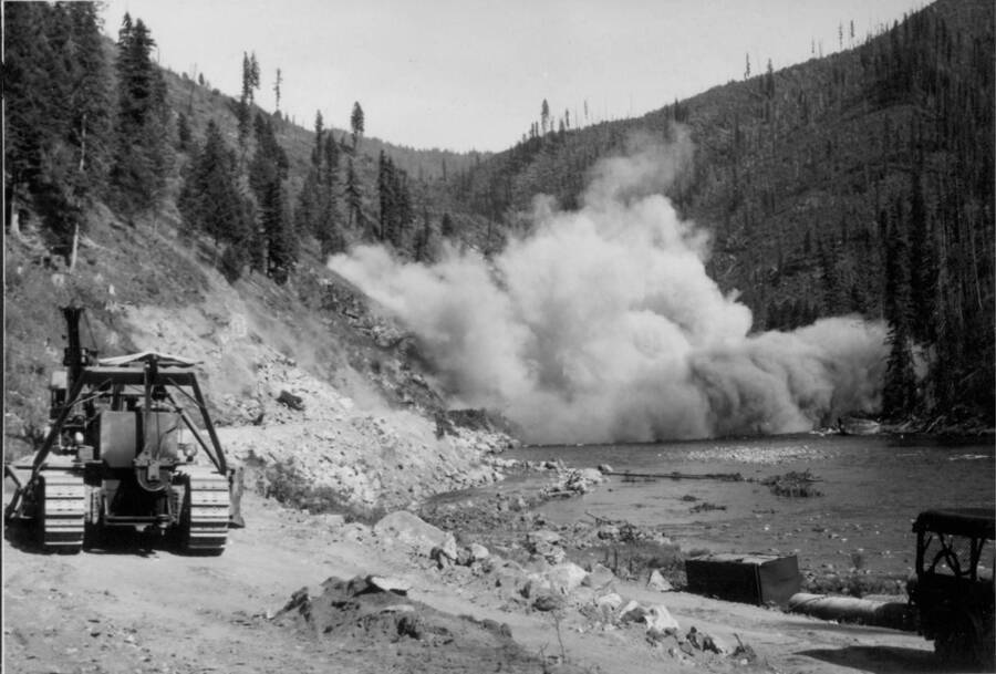 Image of construction equipment by the Lochsa River with explosion in background near the Kooskia Internment Camp. Photo taken from 12-3/4 x 15-1/4 Photograph album of the Kooskia Japanese Internment Camp.