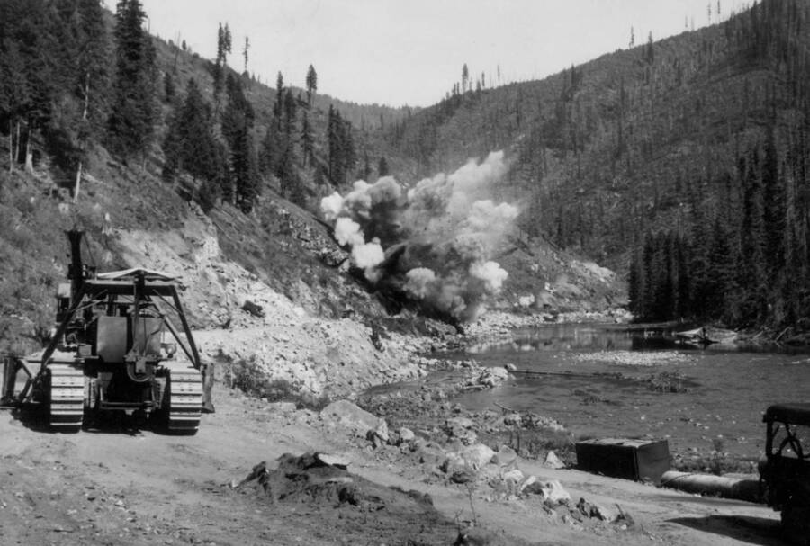 Image of construction equipment by the Lochsa River with a road-blasting explosion in background at Kooskia Internment Camp. Photo taken from 12-3/4 x 15-1/4 Photograph album of the Kooskia Japanese Internment Camp.