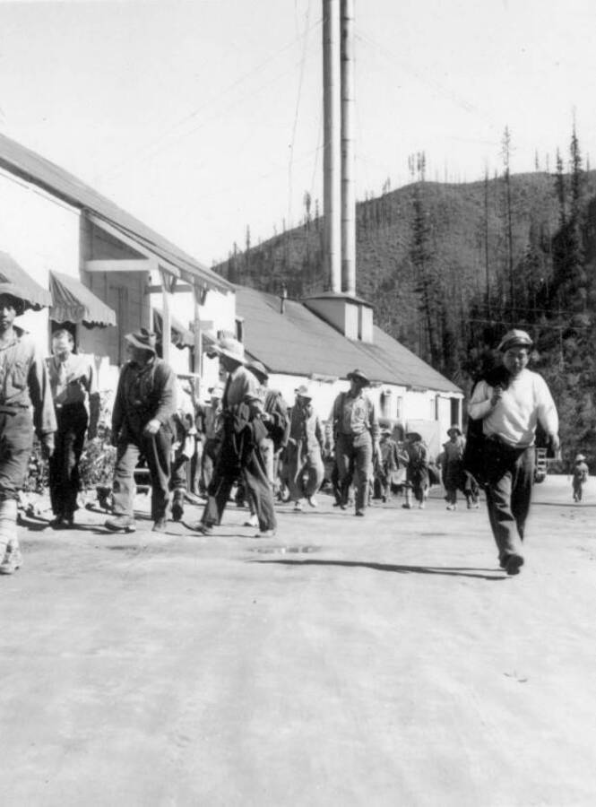Image of people walking in front of the check-in board on the way to a construction project at the Kooskia Internment Camp. Photo taken from 12-3/4 x 15-1/4 Photograph album of the Kooskia Japanese Internment Camp.