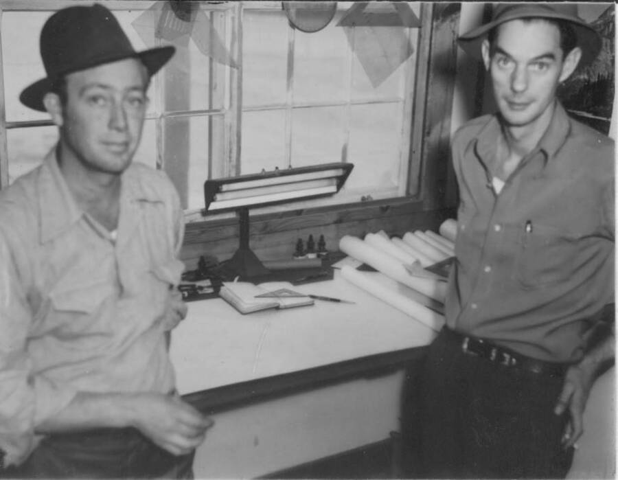 Interior shot of two men inside a building in front of a desk at Kooskia Internment Camp. Drafting and architecture materials can be seen on the desk behind them. Photo taken from 12-3/4 x 15-1/4 Photograph album of the Kooskia Japanese Internment Camp.