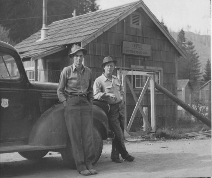 Image of two men leaning on a vehicle in front of the U.S. Public Roads office at Kooskia Internment Camp. Photo taken from 12-3/4 x 15-1/4 Photograph album of the Kooskia Japanese Internment Camp.