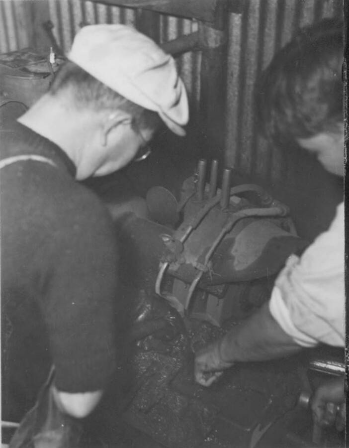 Interior shot  of two men working on an engine in the machine shop at the Kooskia Internment camp. Photo taken from 12-3/4 x 15-1/4 Photograph album of the Kooskia Japanese Internment Camp.