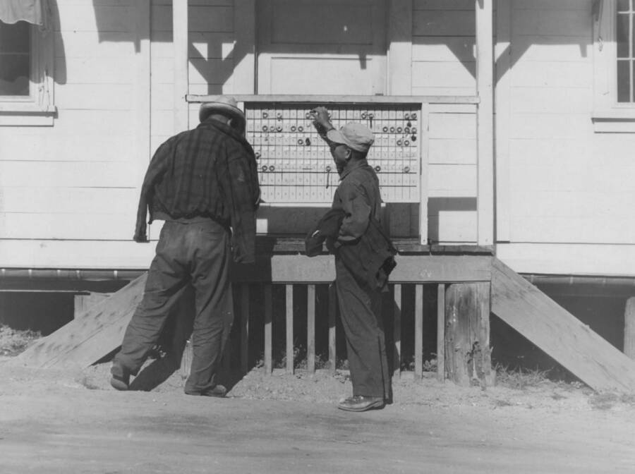 Image of two men outside a building at Kooskia Internment Camp in front of the check-in board. Photo taken from 12-3/4 x 15-1/4 Photograph album of the Kooskia Japanese Internment Camp.