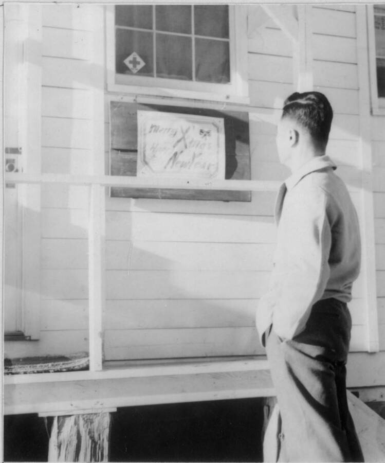 Image of a man looking at a "Merry Christmas, Happy New Year" sign posted outside of a building. Photo taken from 12-3/4 x 15-1/4 Photograph album of the Kooskia Japanese Internment Camp.