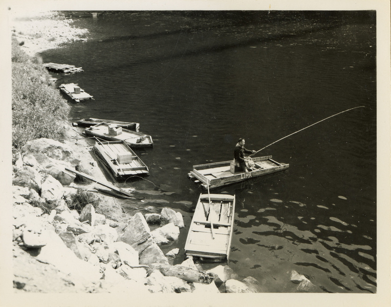 A black and white image of a man fishing near a river. Two boats sit along the bank at the river.