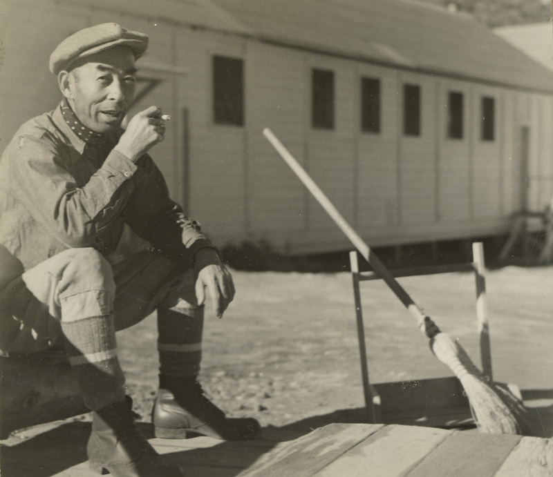 A black and white photo of a man sitting on the steps of a building and smoking a cigarette. A broom leans against a chair to the left of him.