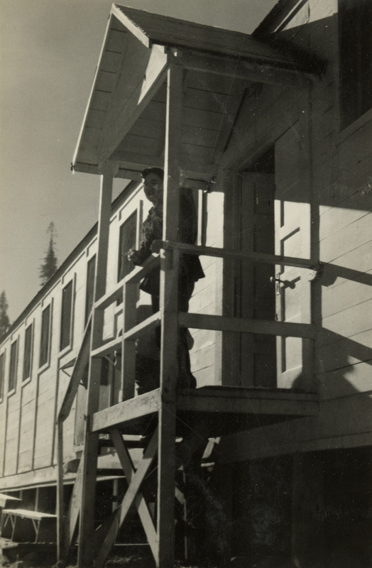 A black and white photograph of a man standing on a high porch of a building. He is leaning along the railin gof the porch.