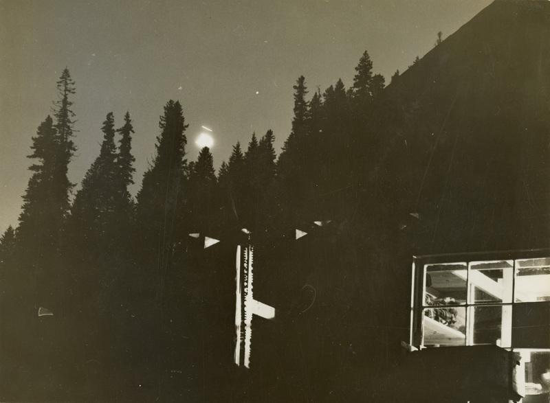 A black and white photograph of the internment camp at night. Lights are shown coming from the windows of a building.