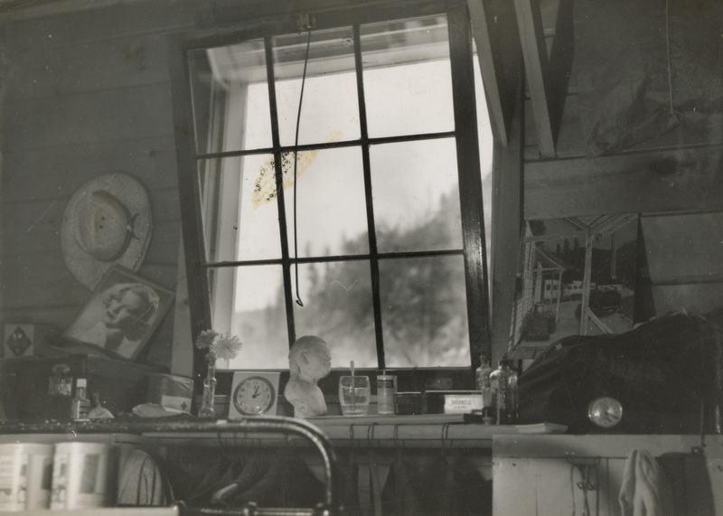 A black and white photograph from the inside of a window. A number of items such as a bust of a woman, glasses, posters, and a clock sit below the window.