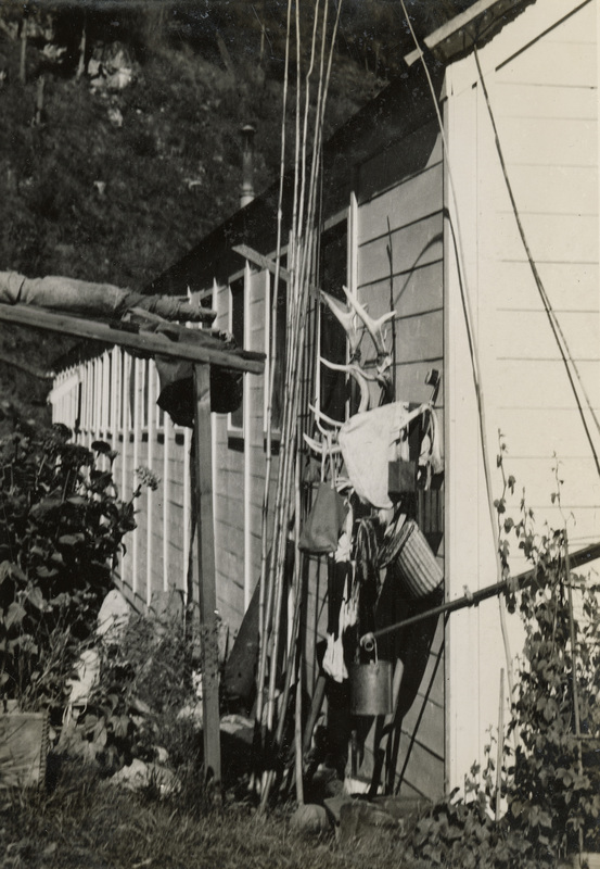A black and white photograph of the outside of a building with fishing and hunting items leaning against the siding of the building. There are plants, grass, and shrubs along the building.