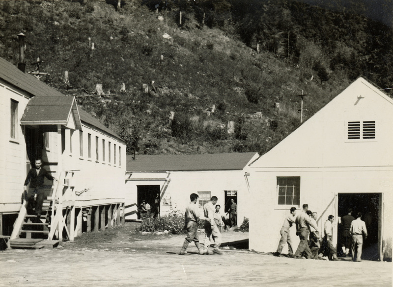 A black and white photograph of men walking outside of buildings at Kooskia. They appear to be walking between two buildings.