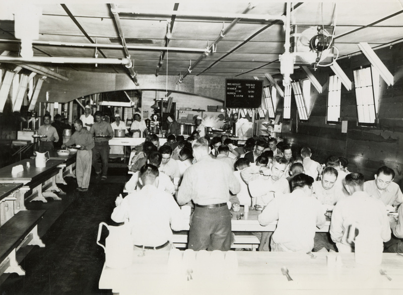 A black and white photograph of men inside of a cafeteria building. THey are eating and interacting with one another.