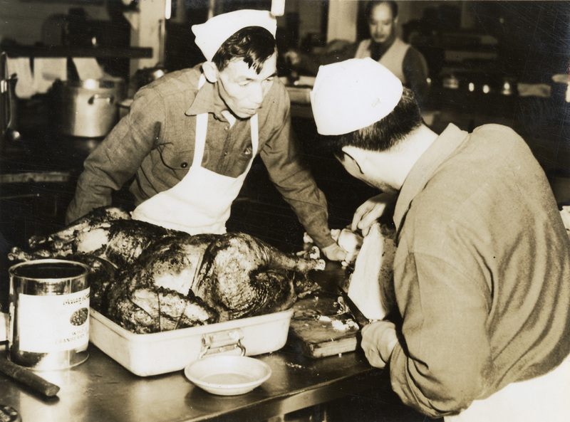 A black and white photograph of two men standing around roasted turkeys. They are carving the turkeys for, assumably, Chirstmas dinner. 