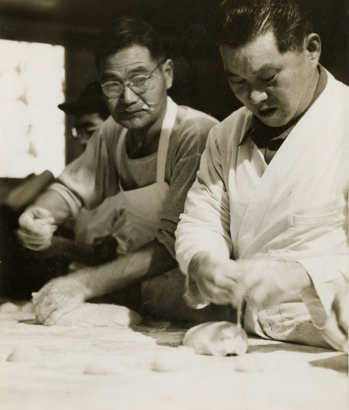 A black and white photograph of two men preparing food. They appear to be making bread.