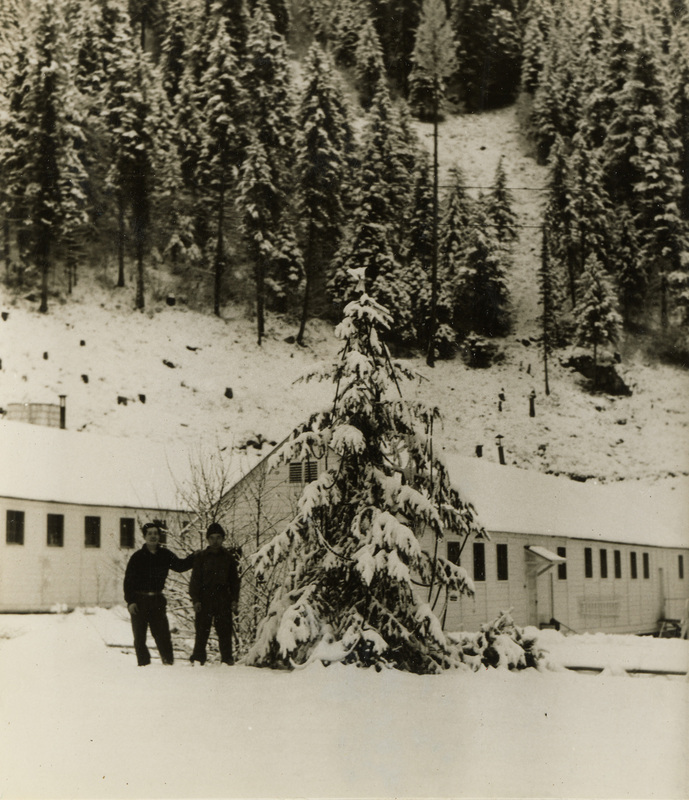 A black and white photograph of two men standing outside in the snow. There is a pine tree in front of the buildings that they are standing next to.