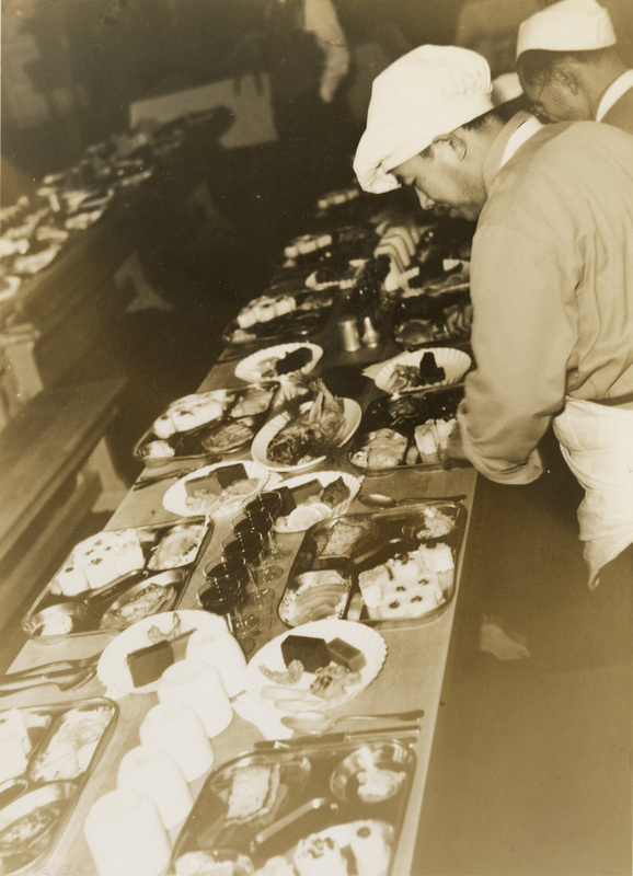 A black and white photograph of men setting a wide array of food around a table.