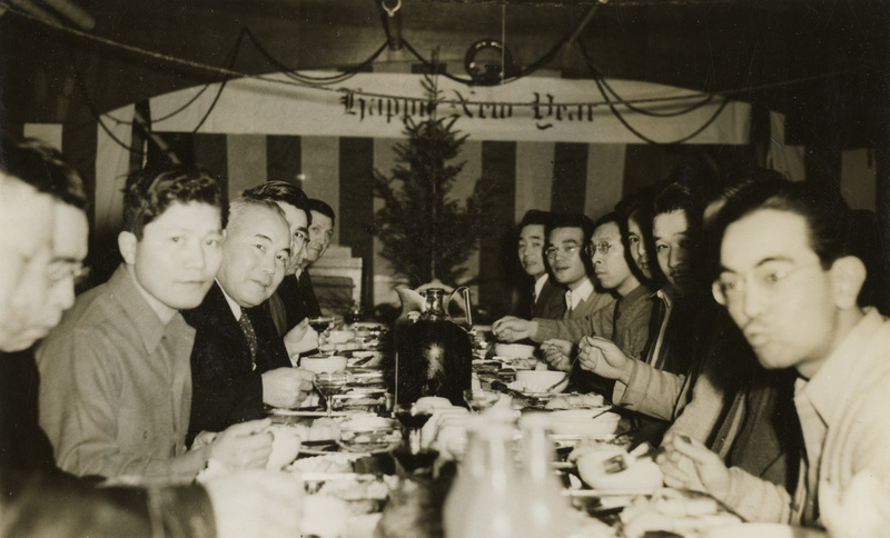 A black and white photograph of men sitting at a table. A white banner in the foreground reads "Happy New Year". The men are eating a dinner.