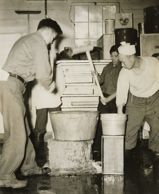 A black and white photograph of four men making mochi. Two men are using hammers to hit the mochi dough, while another man is helping with the dough.