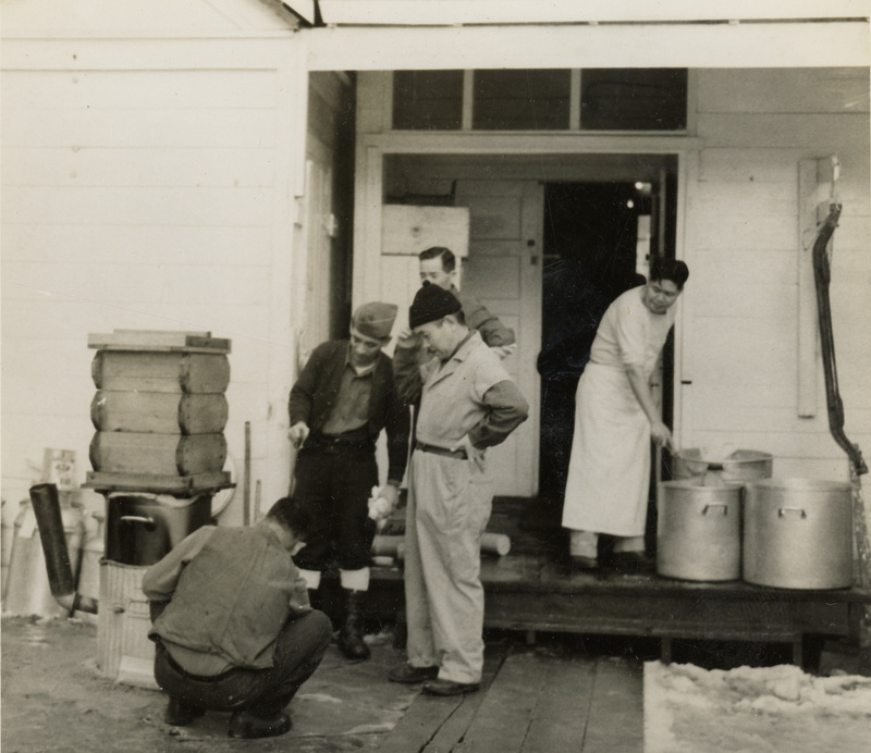 A black and white image of men working on a seiro outside. There are four men looking at the seiro and one man working with a large soup pot.
