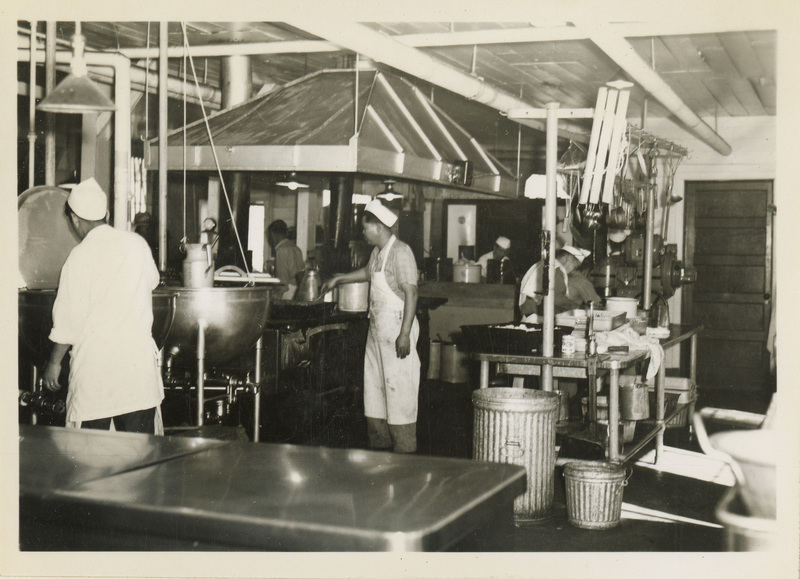 A black and white photo of a kitchen and men working.