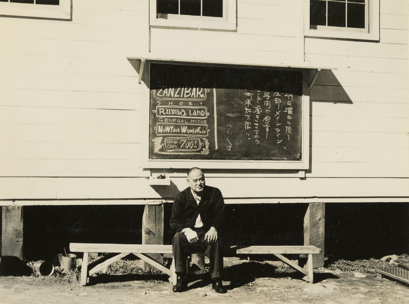 A black and white photograph of a man sitting under a chalkboard on the outside of a building. Both English and Japanese can be read from the sign.