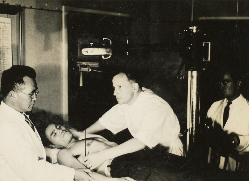 A black and white photograph of a patient on a bed, with three medical personnel attending to the patient on the table. 