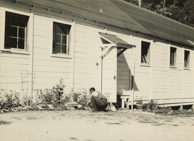 A black and white photo of a man kneeling in front of a flower bed at the base of a building.