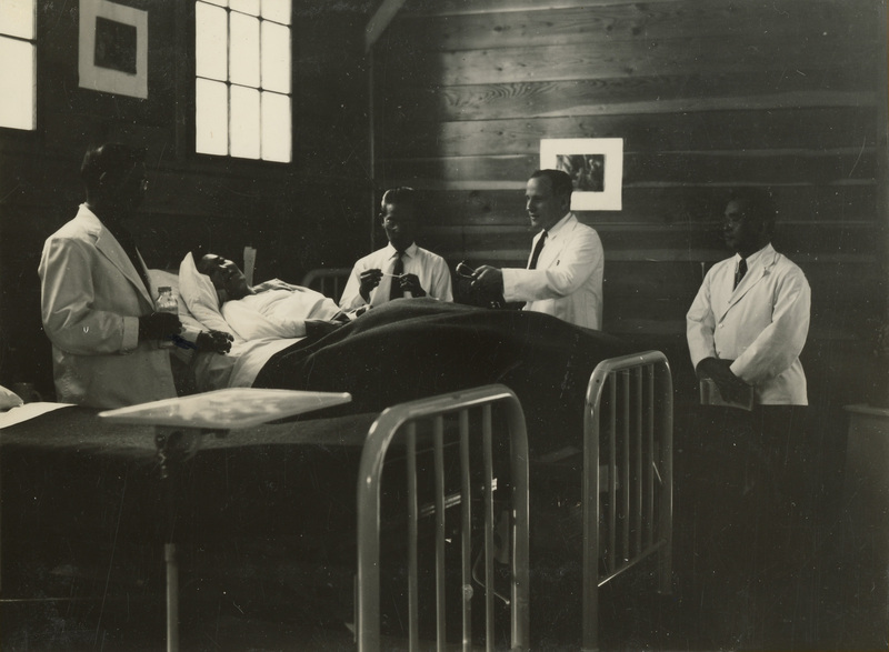 A black and white photo of medical personnel tending to a patient in a room. Four medical personnel surround a man on a bed.