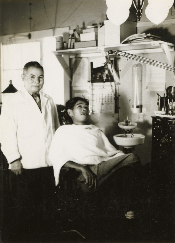 A black and white photograph of a dentist standing next to a dental patient laying back in a chair.