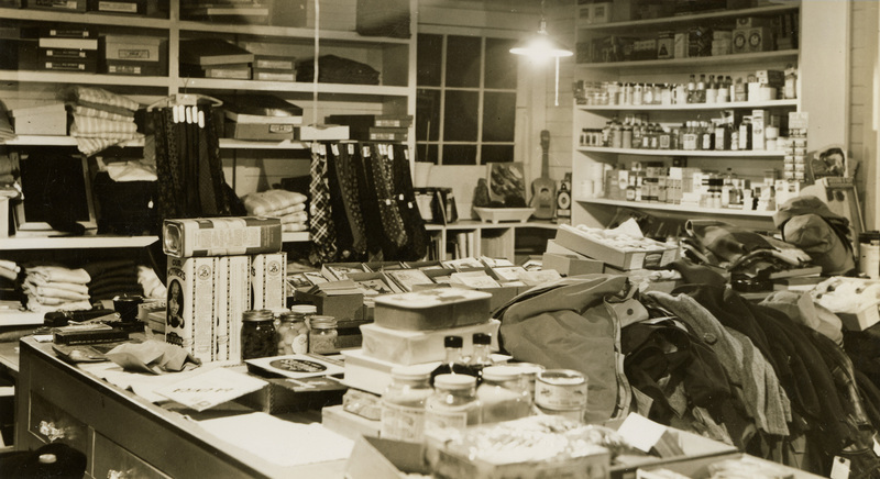 A black and white photograph of the merchandise inside of a store such as bottles, clothing, and food.