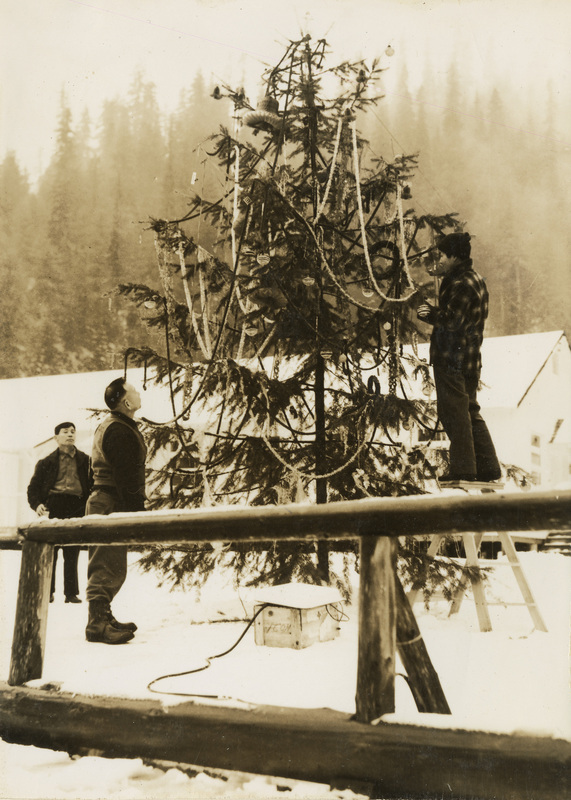 A black and white photograph of three men decorating a pine tree outside in the snow. They are hanging Christmas lights.