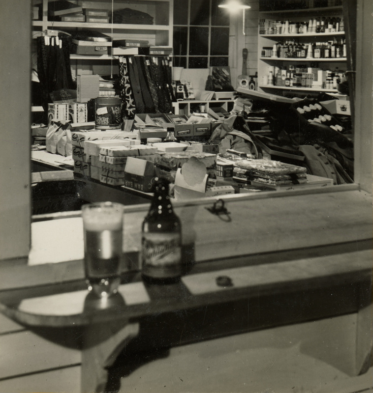 A black and white photograph of the inside of the merchandise room. A beer bottle and beer glass sits in the service window looking into the room.