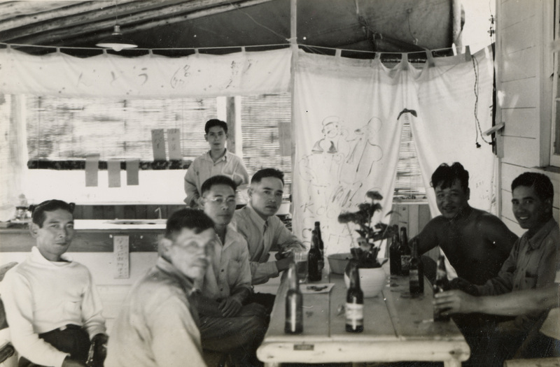 A black and white photograph of seven men sitting at a table on a covered patio. On the table are some bottles.