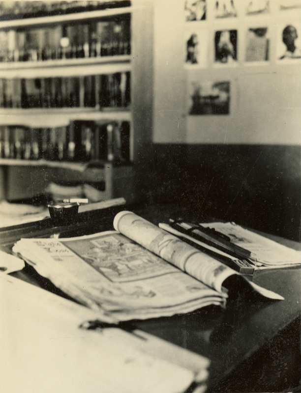 A black and white photograph of a magazine open on a table. A large bookshelf is in the background.