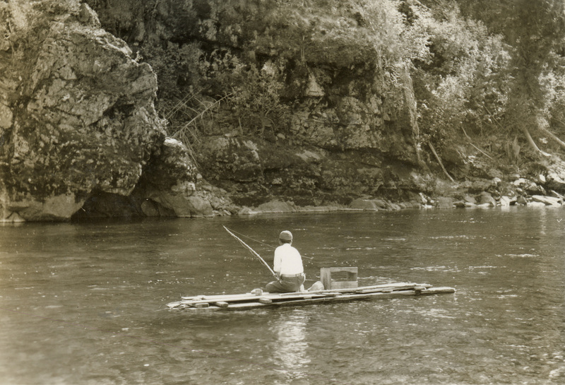 A black and white photograph of a man fishing on a raft in a river.