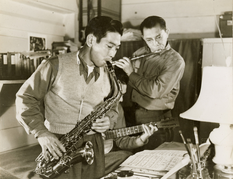 A black and white photograph of three men playing instruments: one playing a saxophone, one playing a guitar, and one playing a flute. 