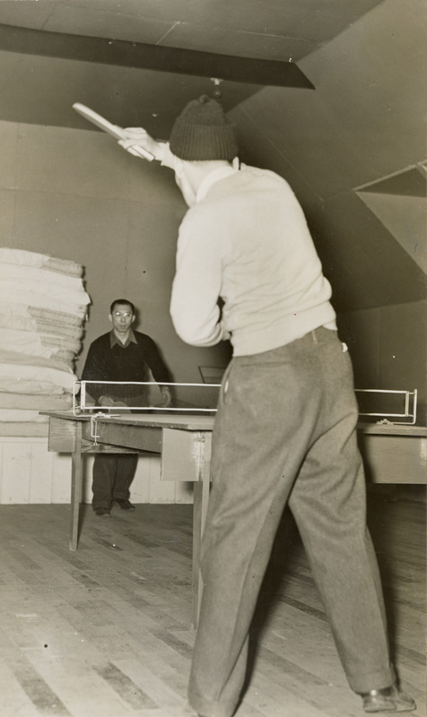 A black and white photo of two men playing table tennis, also known as ping pong, in a building. One man has his back to the camera. 
